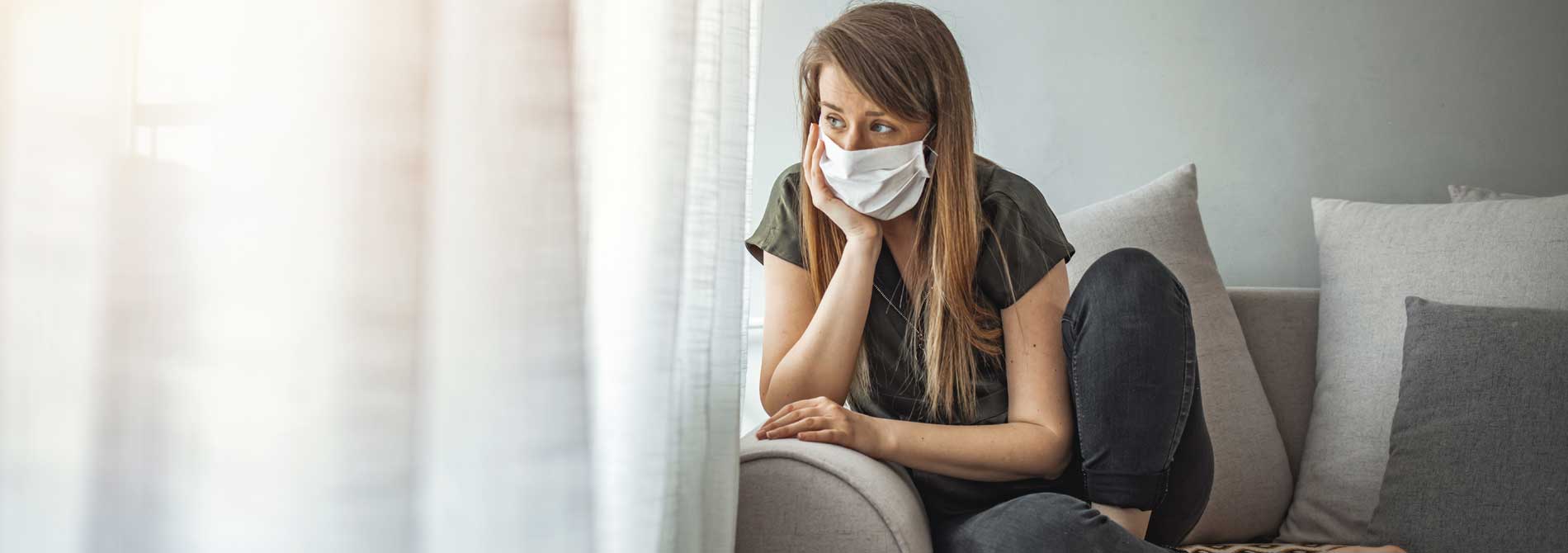 woman indoors wearing surgical mask looking out window